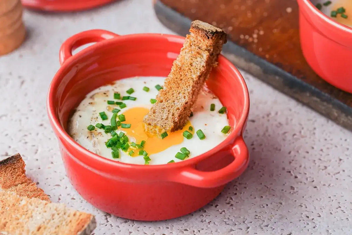 Un œuf cocotte appétissant dans un ramequin rouge, garni de ciboule et d'une tranche de pain grillé.