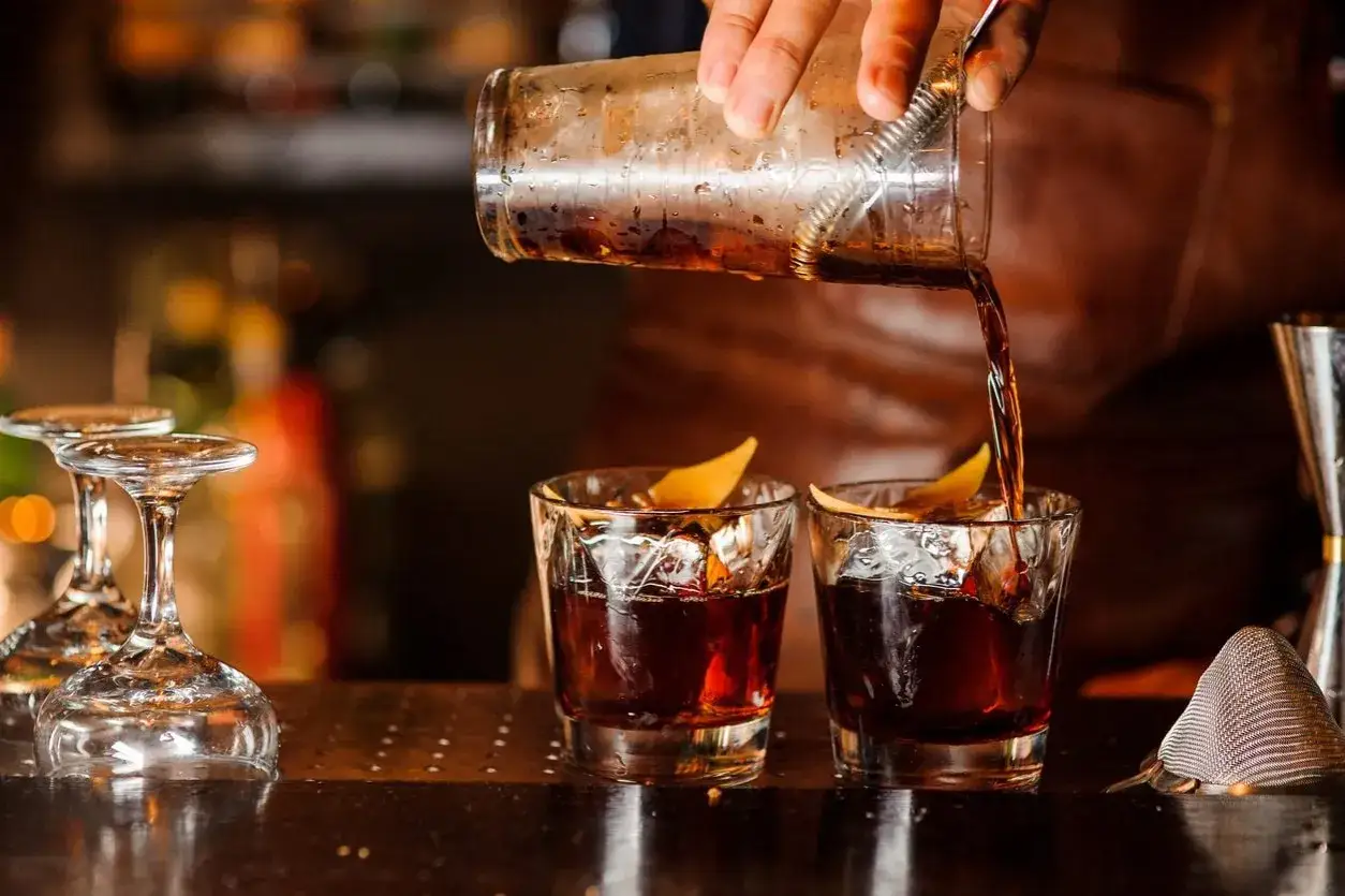 Un barista versa un **bitter cocktail** in due bicchieri con ghiaccio e scorza d'arancia.