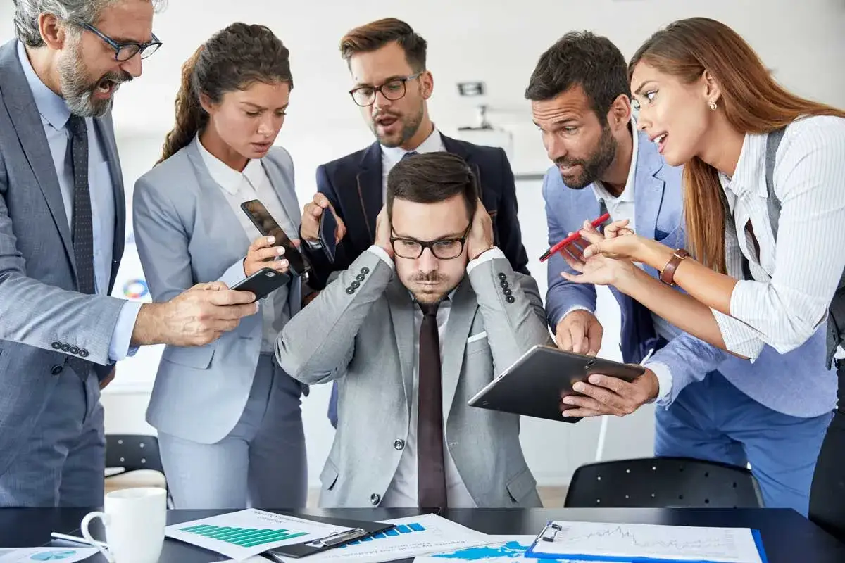 Ein gestresster Mann mit Händen an den Ohren, umgeben von Kollegen, die ihn mit Handys und Tablets bedrängen. Die Szene vermittelt starke **Angst vor der Arbeit**.