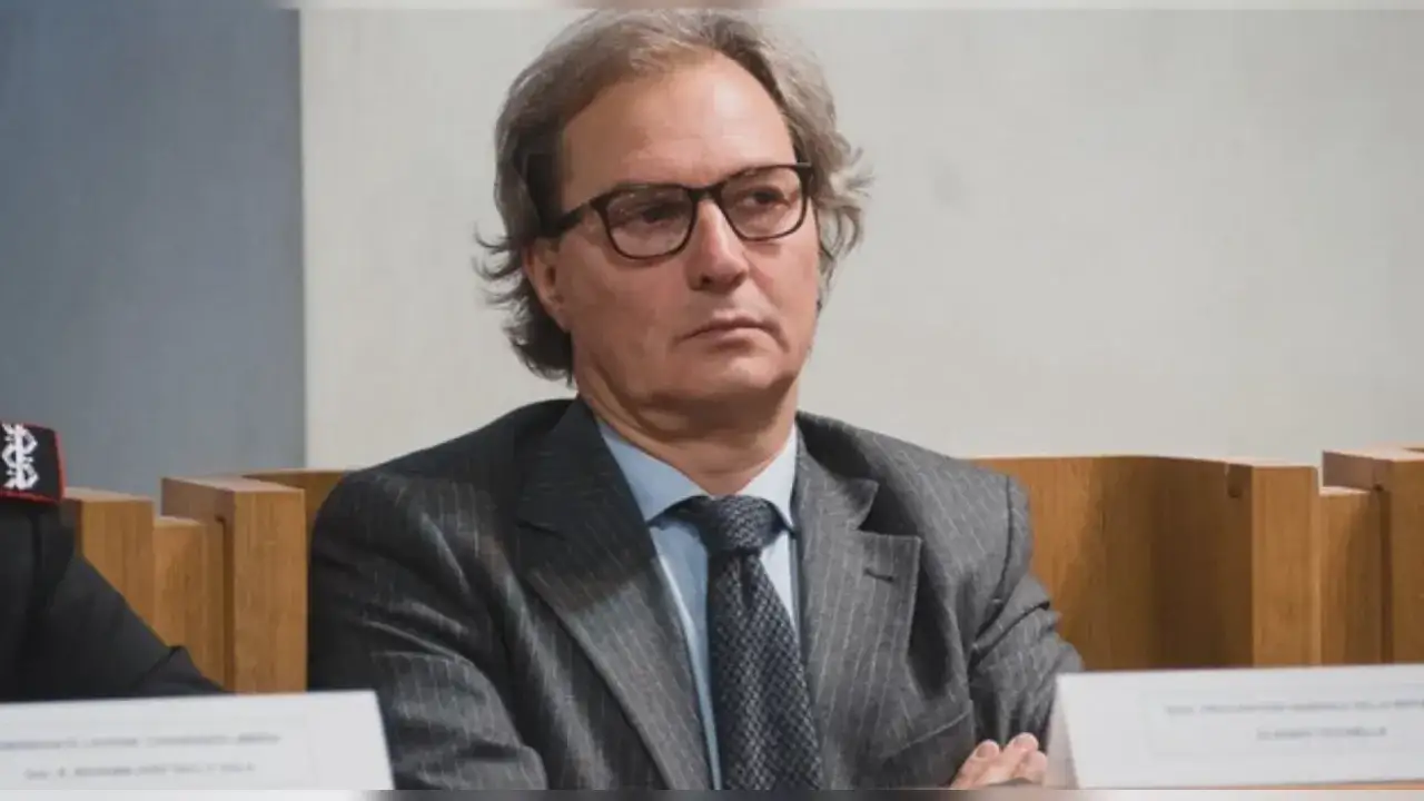 Uomo con occhiali e abito a righe, seduto a un tavolo. Si discute di cosa fa un procuratore.