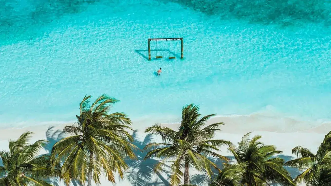 Traumhafter November auf den Malediven: Ein Mann watet durch kristallklares Wasser zu einer Schaukelinsel. Palmen s&auml;umen den wei&szlig;en Sandstrand.