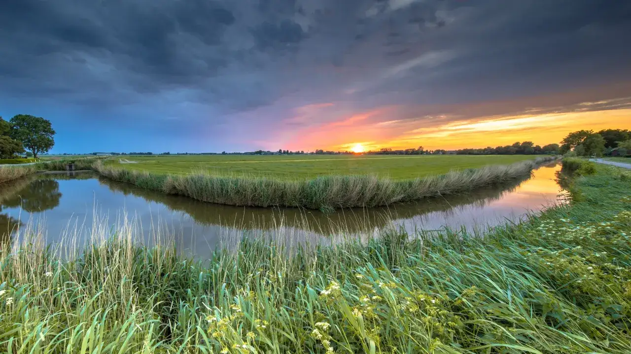 Coucher de soleil spectaculaire sur un paysage rural hollandais, capturé en photos grand angle. Un canal serpente à travers des champs verdoyants, reflétant le ciel aux teintes orangées et bleues.