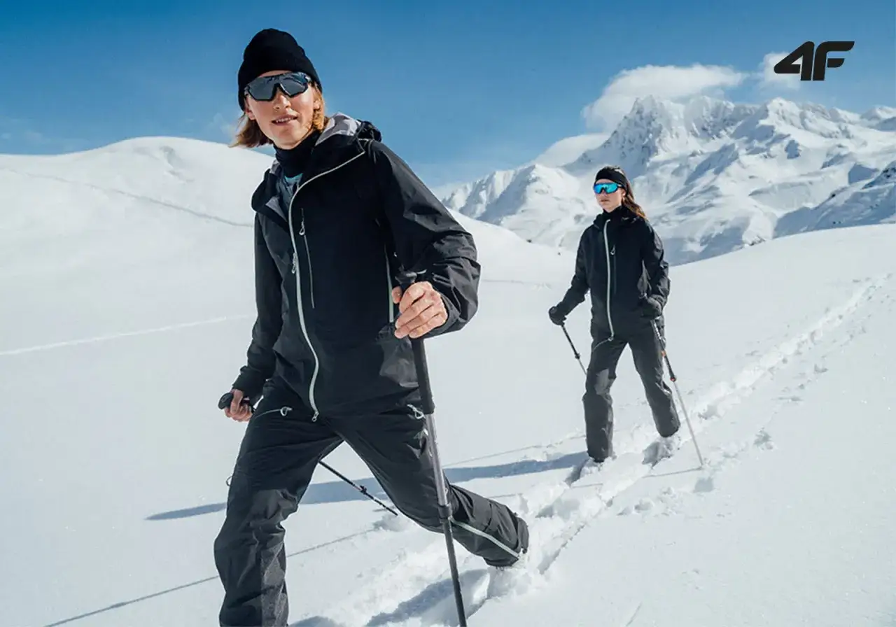 Dwóch narciarzy w czarnych strojach przemierza ośnieżone szczyty. Ich ski tury prowadzą przez dziewiczy puch, a słońce odbija się w goglach.