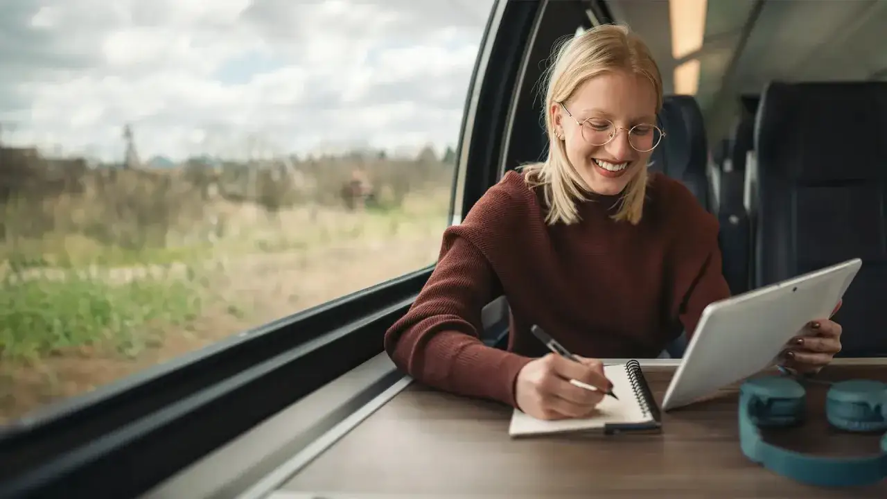Junge Frau im Zug nutzt Tablet und Notizbuch, um ihre Zeit effizient zu gestalten. Perfekt f&uuml;r IU Teilzeit 1 Zeitaufwand.