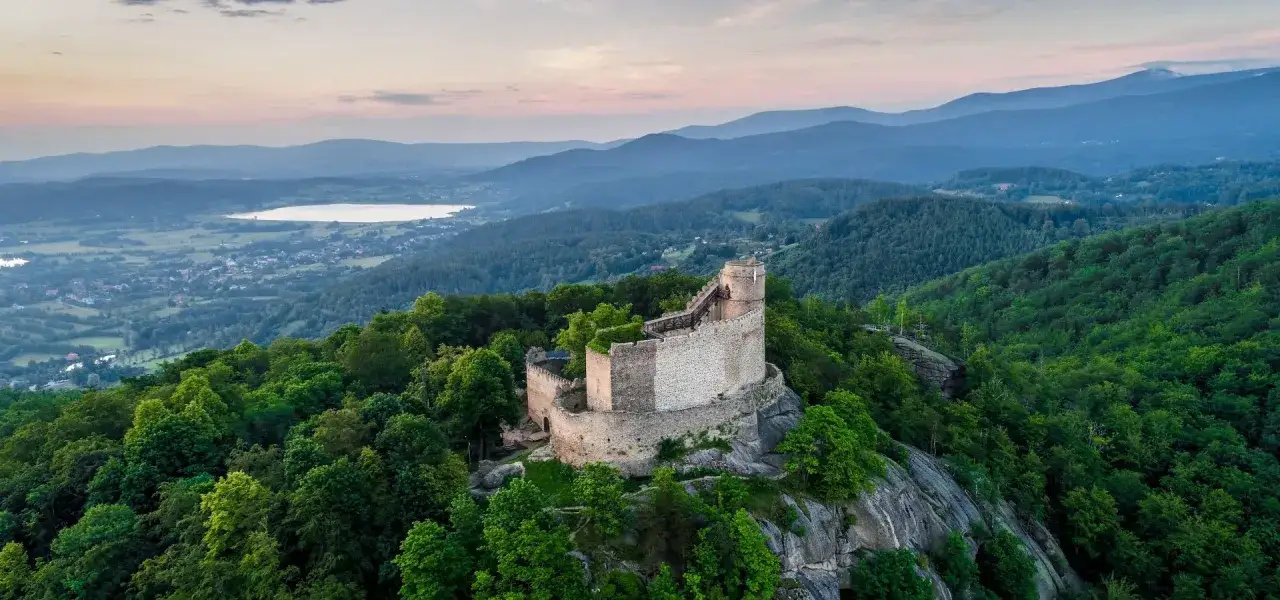 Zamek Chojnik góruje nad zielonymi wzgórzami i jeziorem. Ruiny zamku, otoczone drzewami, wyglądają jak z bajki.