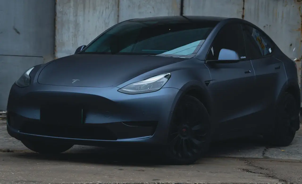 Mörkgrå Tesla Model Y med matt finish, som svart vax, står framför en sliten betongvägg.