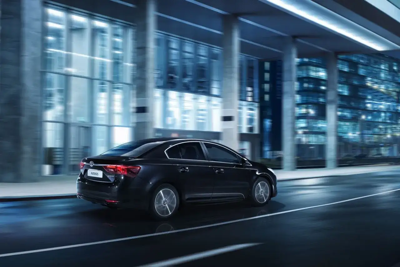 Ciemny sedan Toyota Avensis T27 1.8 benzyna jedzie nocą po mieście, mijając nowoczesne budynki.