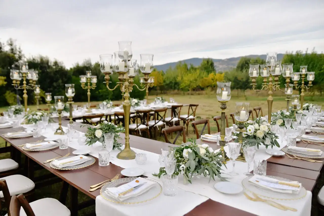 Table dressée pour un mariage en extérieur, avec des chandeliers dorés, des fleurs blanches et des bougies allumées. La date de mariage est inscrite sur les menus.