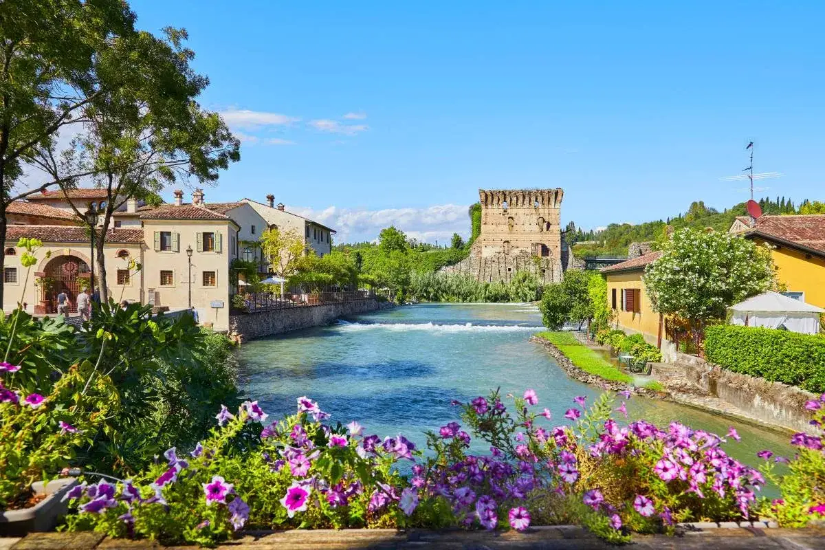 Un incantevole borgo da visitare in Veneto, con un fiume che scorre tra case antiche e una torre medievale, circondato da fiori colorati.