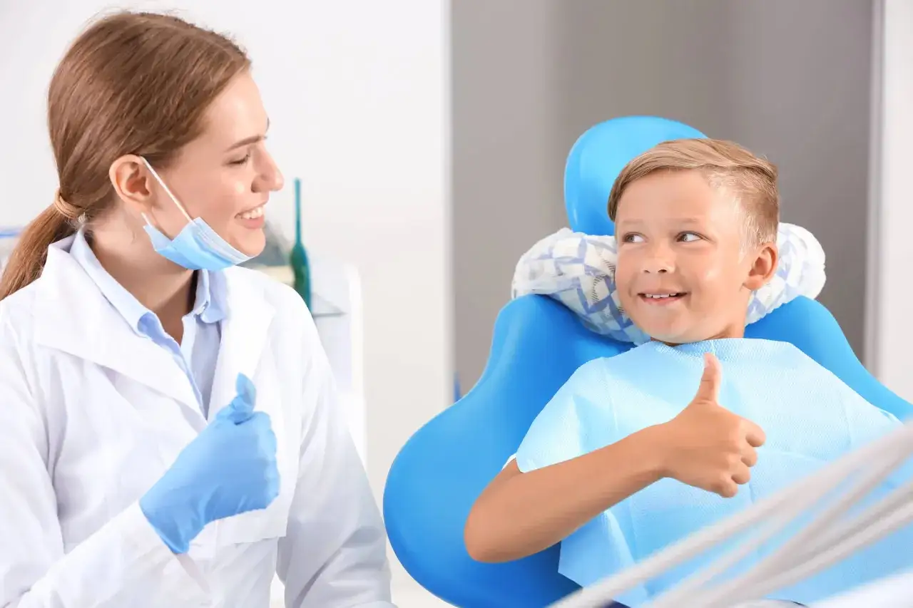 Uśmiechnięty chłopiec po zabiegu lakowania zębów daje kciuk w górę. Dentystka w białym fartuchu i rękawiczkach również pokazuje kciuk w górę.