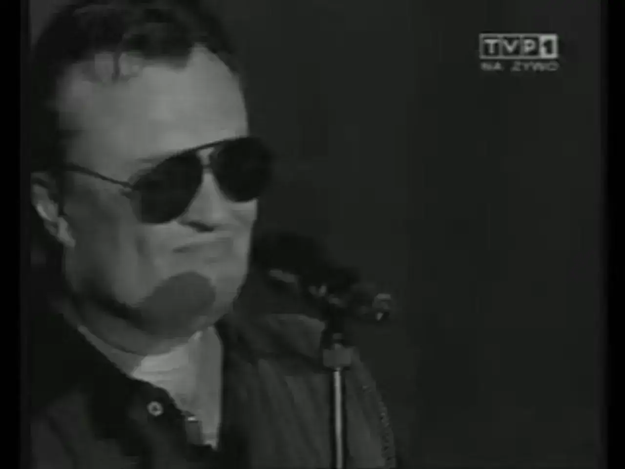 Mężczyzna w okularach przeciwsłonecznych śpiewa do mikrofonu. W tle logo TVP1. Jakby był w 