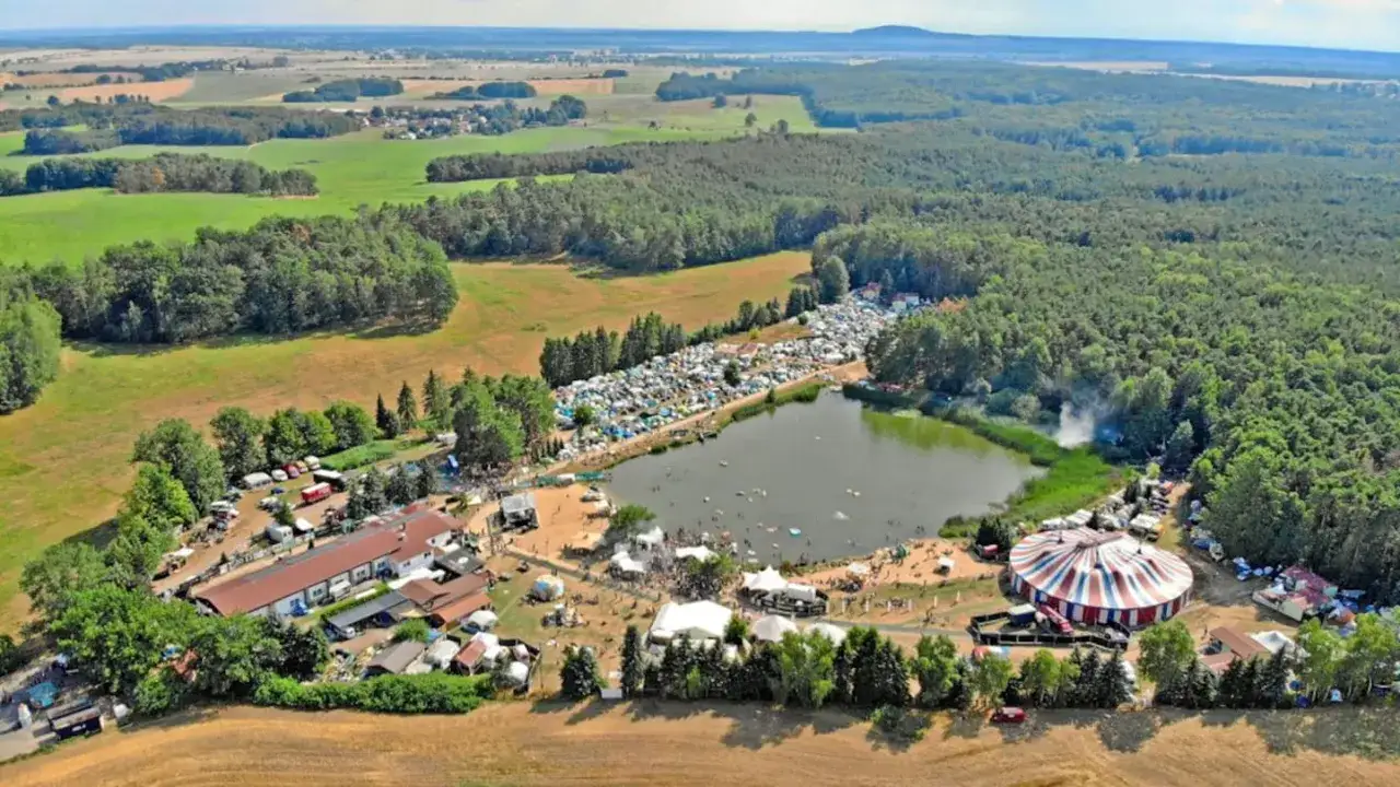 Luftaufnahme eines großen Festivals am See mit Zelten, Bühnen und einem Zirkuszelt. Das Nachtigall Festival ist in vollem Gange.