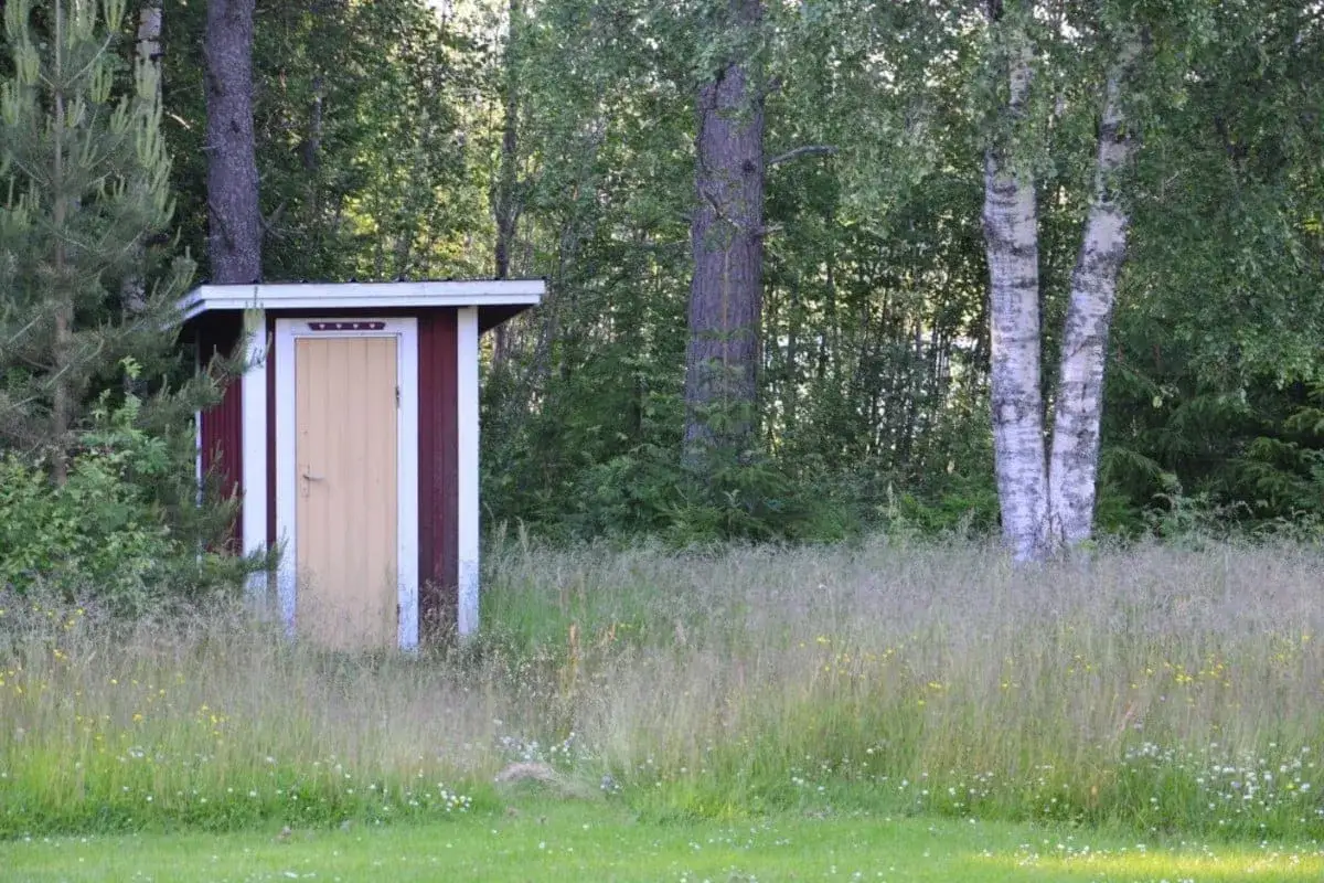 Ett r&ouml;tt utedass med gul d&ouml;rr st&aring;r i en &auml;ng med h&ouml;gt gr&auml;s och blommor, omgivet av skog. Perfekt f&ouml;r att bygga utedass.