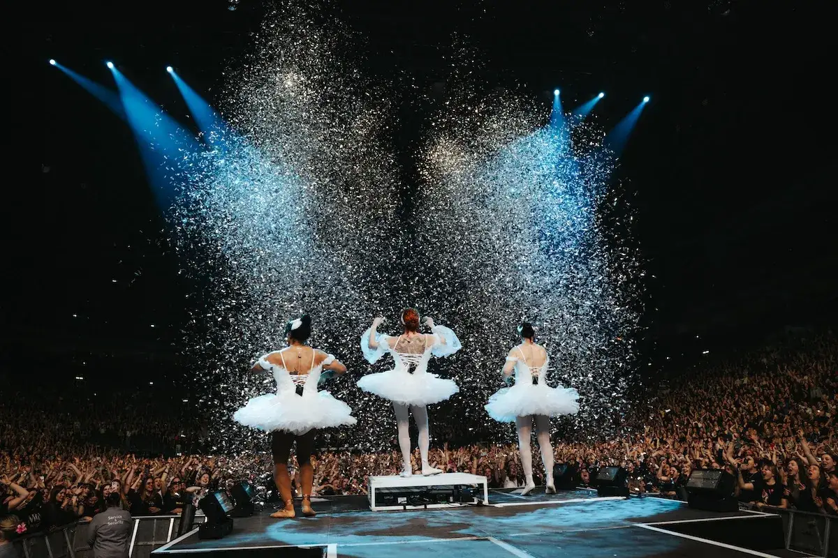Drei T&auml;nzerinnen in wei&szlig;en Tutus stehen auf einer B&uuml;hne, w&auml;hrend Konfetti f&auml;llt. Ein Meer von Fans jubelt. Ein Moment des puren kiz frieden.