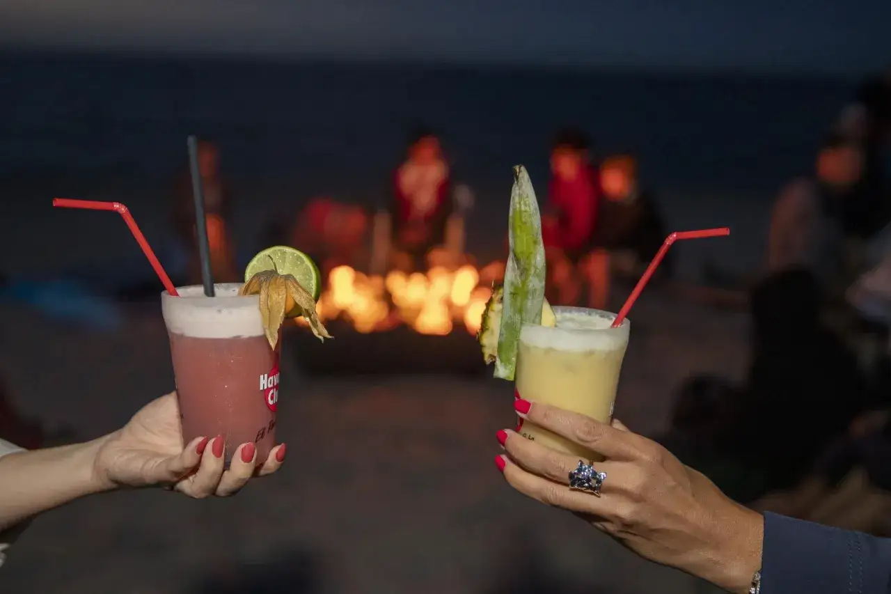 Zwei Cocktails am Strand bei Nacht. Im Hintergrund ein Lagerfeuer und verschwommene Silhouetten von Menschen. Perfekt für eine Ostsee Party.