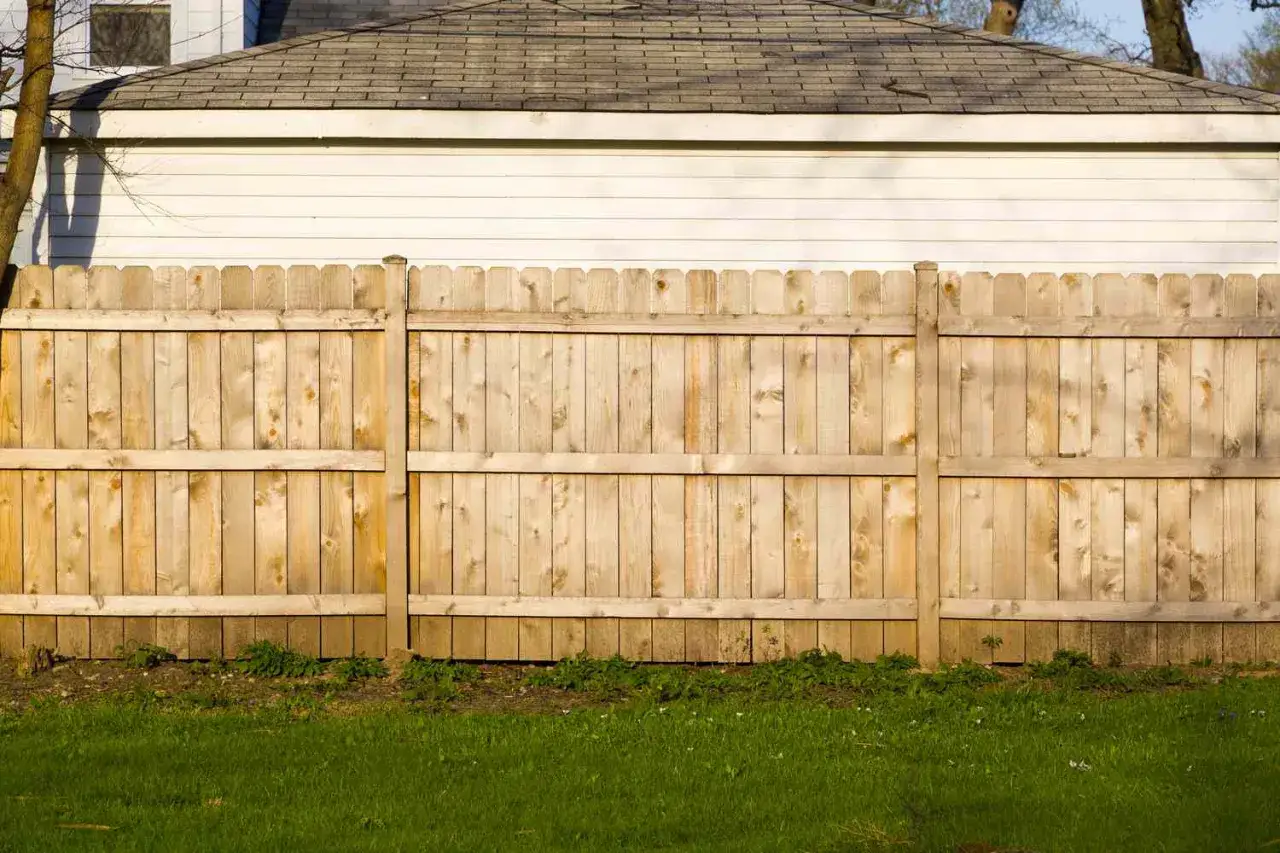 A light brown, wooden fence with a scalloped top, likely made from DIY fence panels, stands before a white garage.