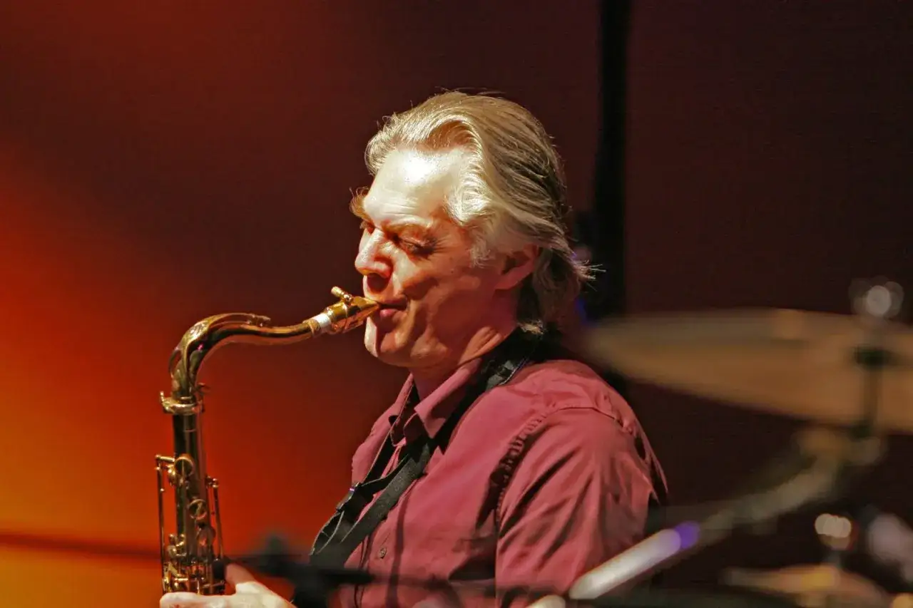 Jan Garbarek, z siwymi włosami i w bordowej koszuli, gra na saksofonie tenorowym.