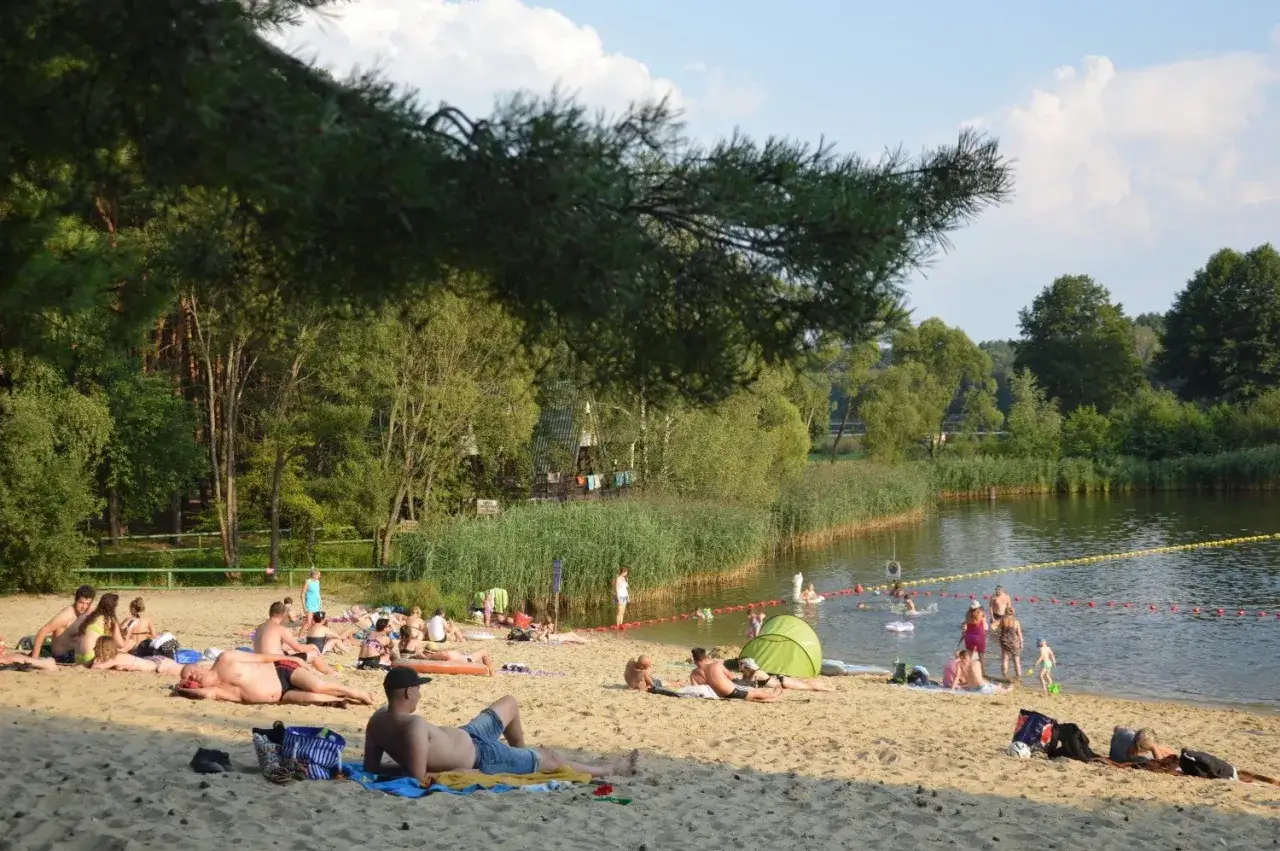 Ludzie odpoczywają na piaszczystej plaży nad jeziorem w Bełchatowie. Kąpielisko tętni życiem, w wodzie pluskają się dzieci i dorośli.