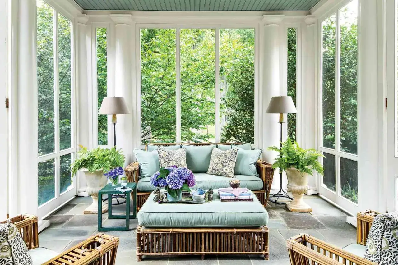 A serene screened-in patio with light blue cushions on rattan furniture, surrounded by lush greenery visible through large windows.
