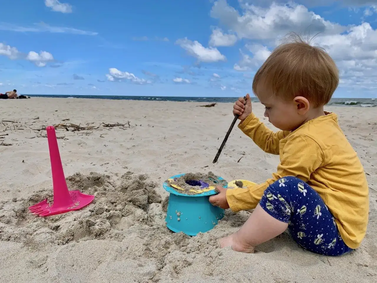 Małe dziecko w ż&oacute;łtej bluzce i granatowych legginsach z pszcz&oacute;łkami bawi się patykiem w piasku na plaży. Obok wiaderko i r&oacute;żowa łopatka.