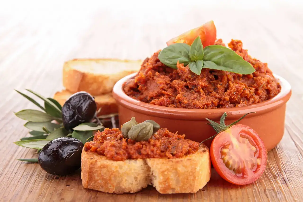 Pyszna pasta z suszonych pomidorów na grzankach, ozdobiona kaparami, oliwkami i bazylią. Idealna na przekąskę.