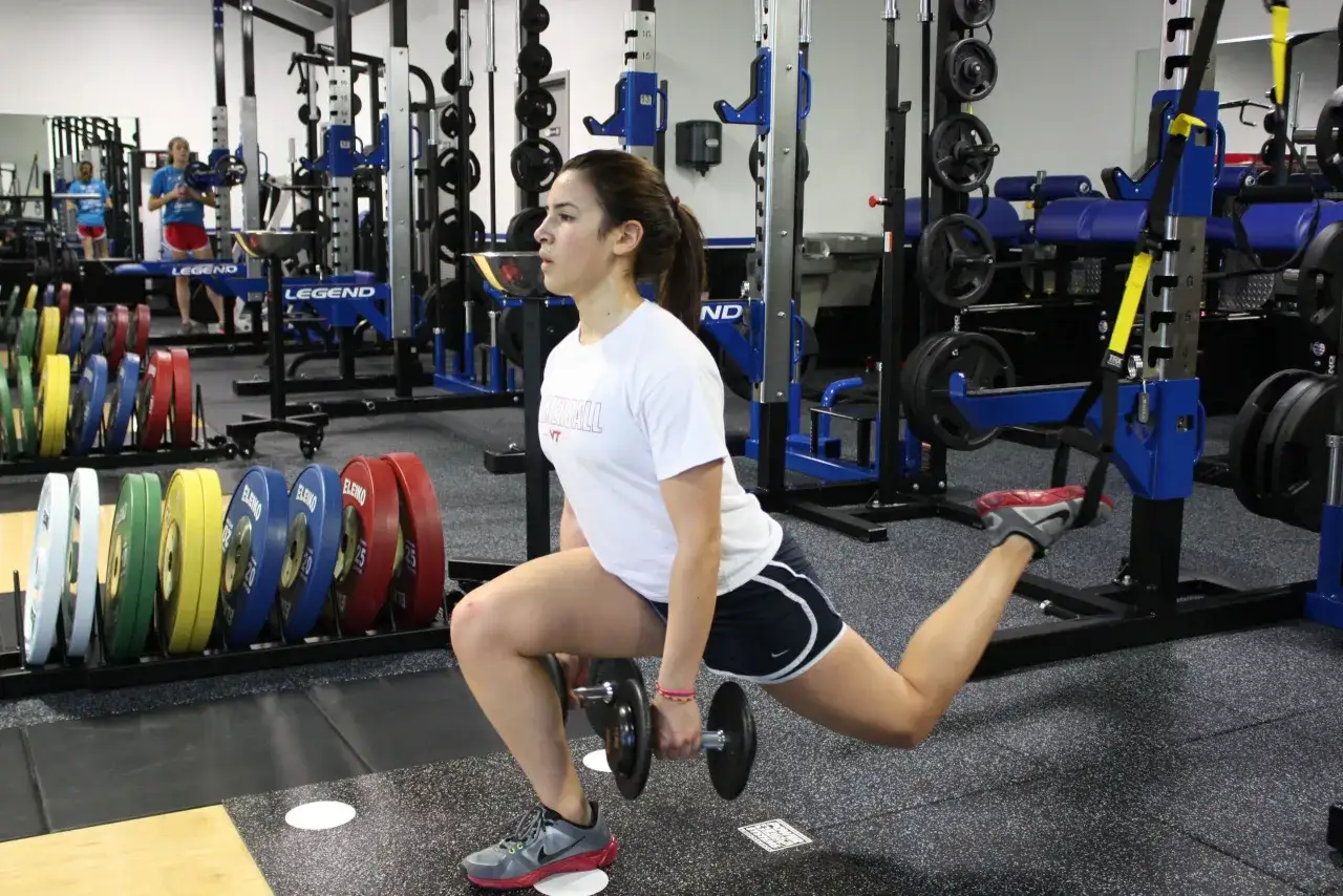 Mujer haciendo zancadas búlgaras con mancuernas en un gimnasio, entrenando como Mijailo Mudryk.