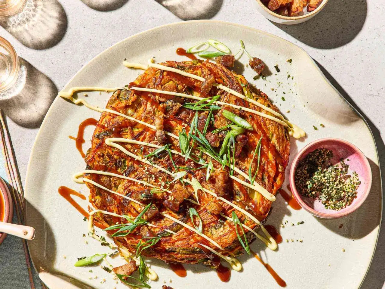 En gyllene okonomiyaki toppad med strimlad salladslök, krispigt fläsk och ringlad okonomiyaki sås samt majonnäs.