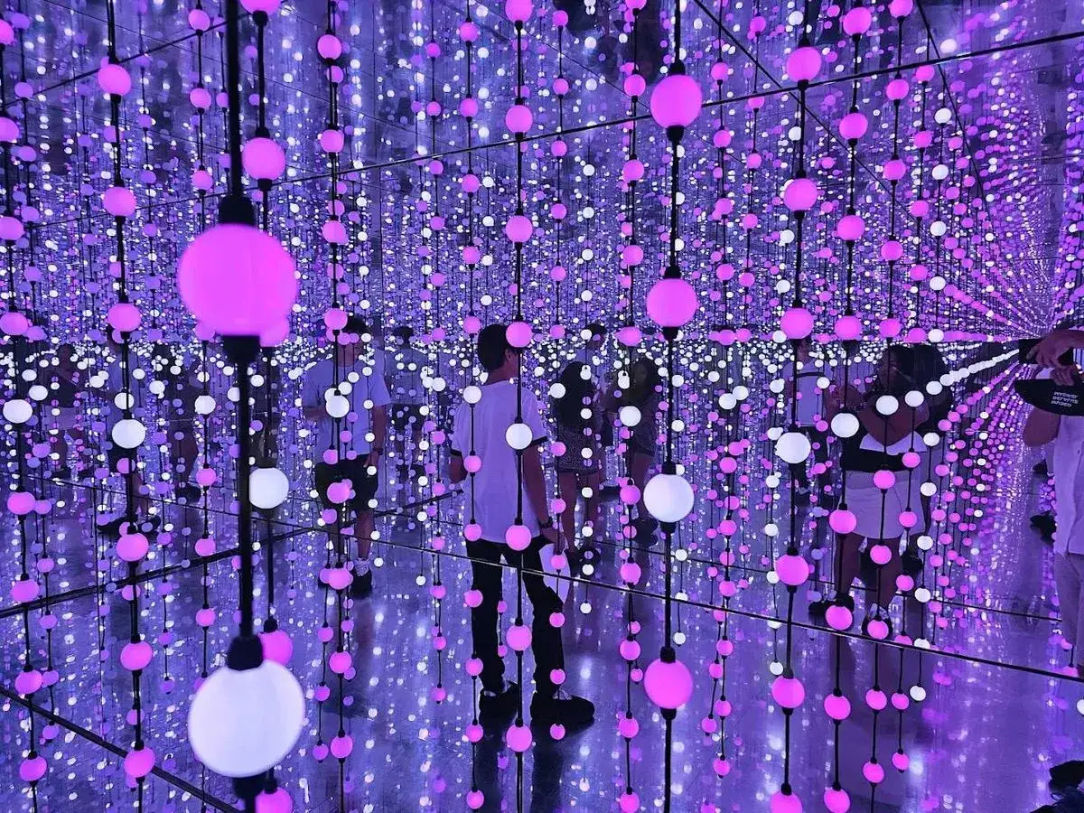 Gente explorando un **nomad museo inmersivo** lleno de luces colgantes rosas y blancas, creando un efecto infinito.