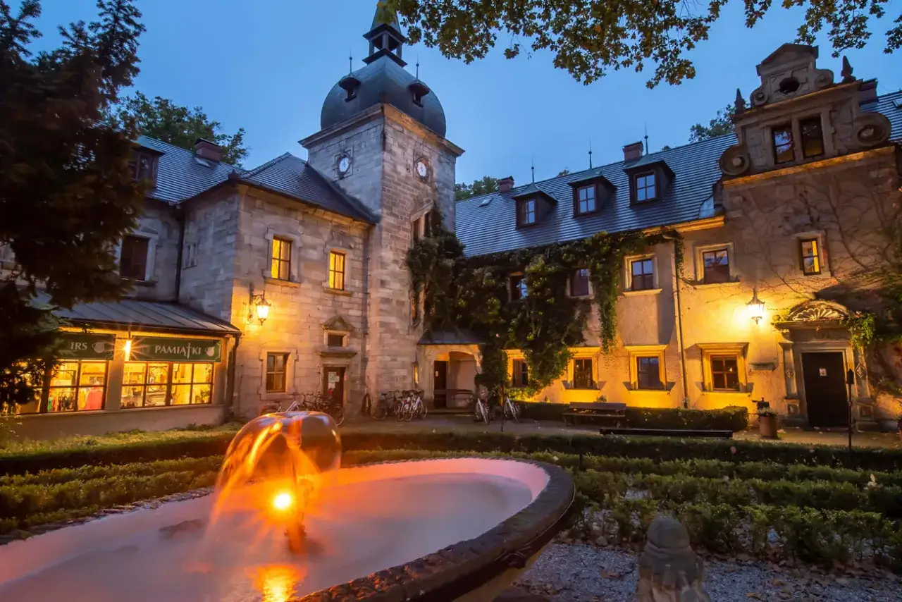 Zamek Kliczków nocą, z fontanną w pierwszym planie i oświetlonymi oknami.