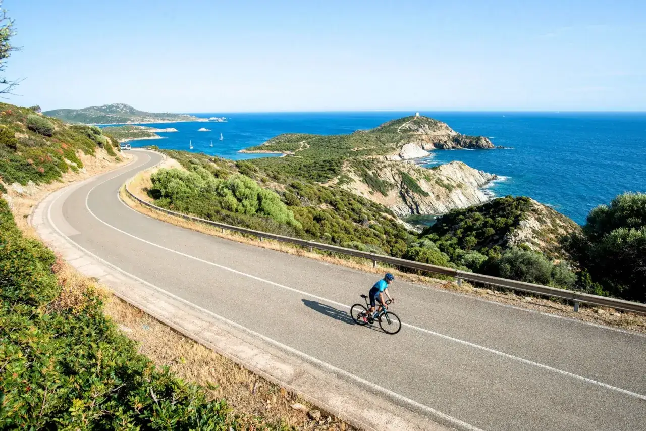 A cyclist enjoys a scenic bike tour in Sardinia, riding along a winding coastal road with the turquoise sea and rugged cliffs in the background.