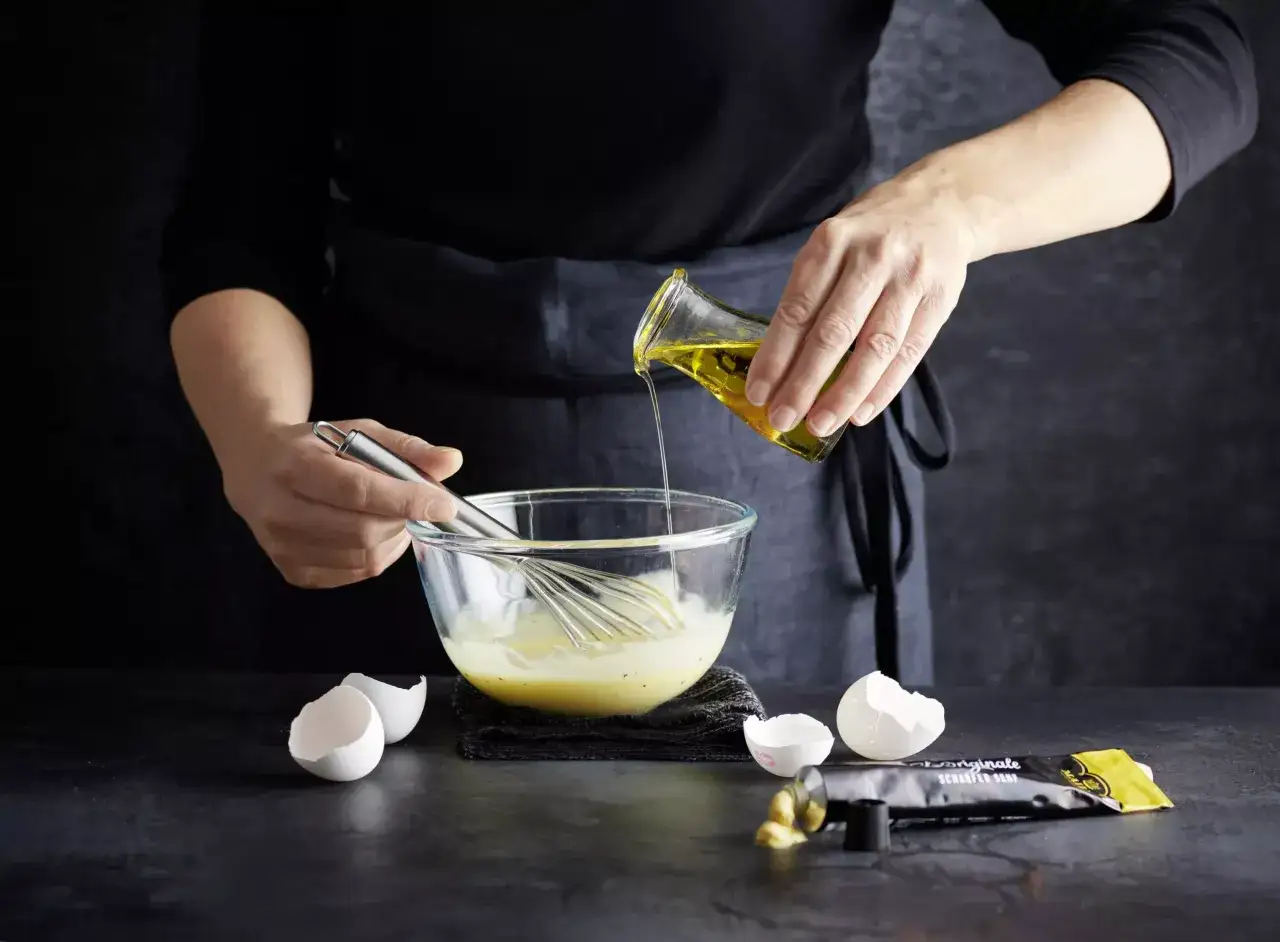 Préparation d'une mayonnaise maison : de l'huile d'olive est versée dans un bol contenant des œufs battus et fouettés.