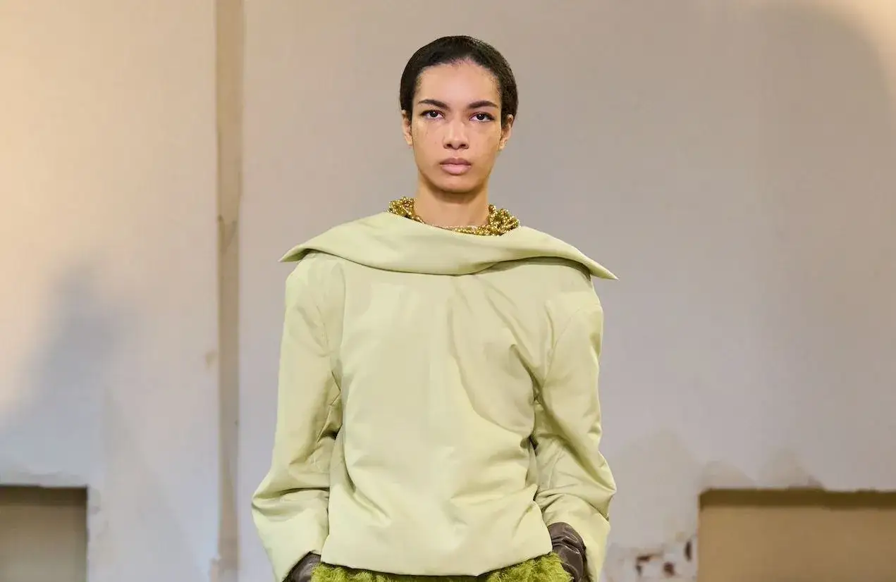 Mannequin portant un haut ample couleur matcha, col drap&eacute;, collier dor&eacute; audacieux et gants marron.
