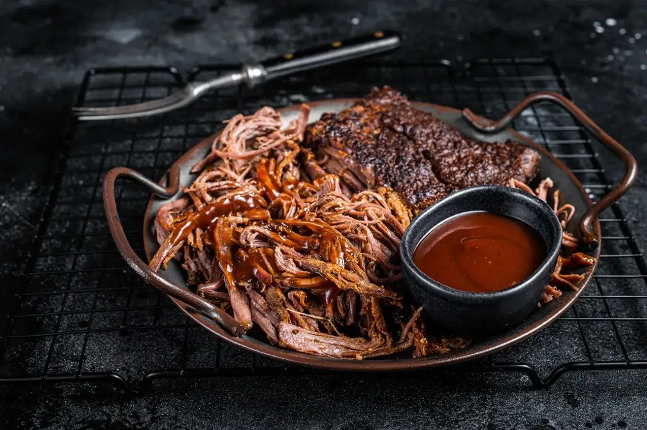 Saftiges pulled pork mit BBQ-Sauce und einem Stück Brisket, serviert auf einem Teller mit Beilagen. Ein Gabel liegt daneben.