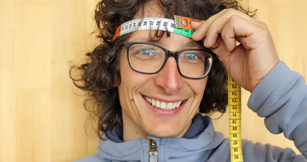 Uomo con occhiali e capelli ricci sorride mentre usa un metro per capire come misurare la circonferenza della testa.
