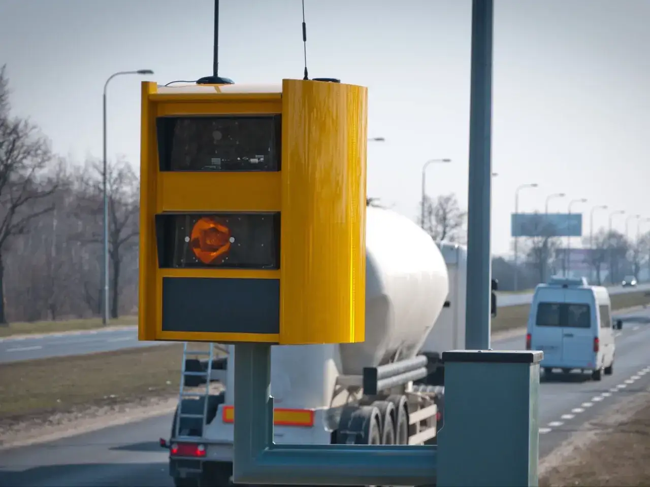 Żółty fotoradar na poboczu drogi, gotowy do działania, gdy przychodzi mandat z fotoradaru. W tle ciężarówka i biały bus.