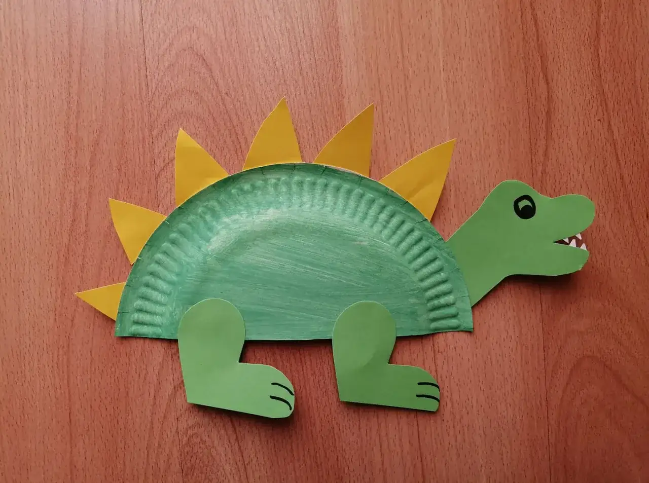 Zielony dinozaur z papierowego talerzyka, ozdobiony żółtymi kolcami. Urocza dinozaur praca plastyczna.