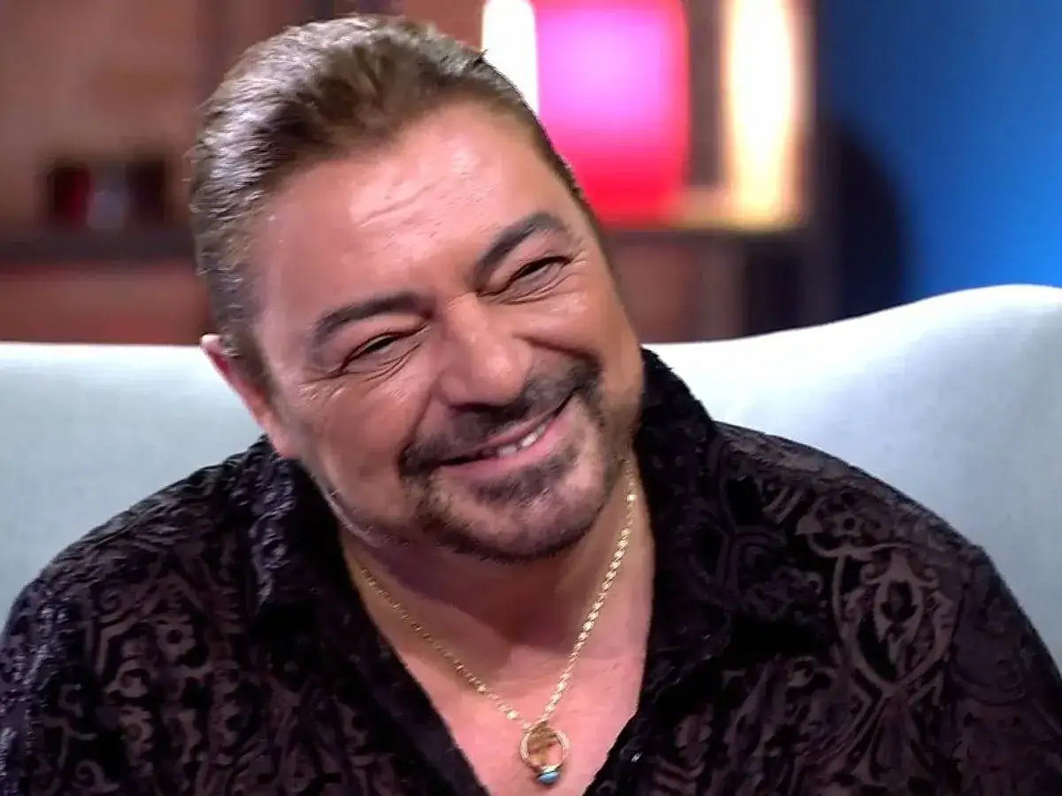 Antonio Canales bailaor, con una sonrisa p&iacute;cara y un collar dorado, luce una camisa negra con un estampado intrincado.