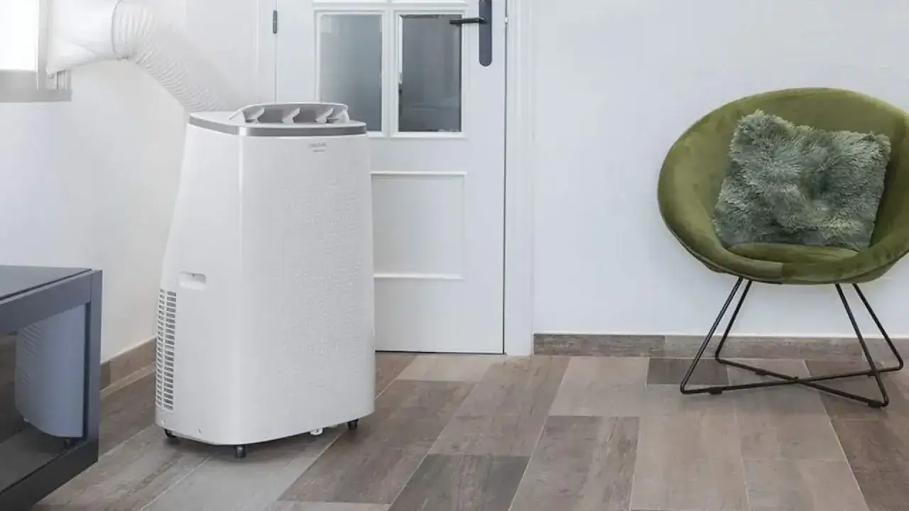 Un climatizador evaporativo port&aacute;til con tubo de extracci&oacute;n conectado a la ventana, junto a una silla verde y una puerta blanca.