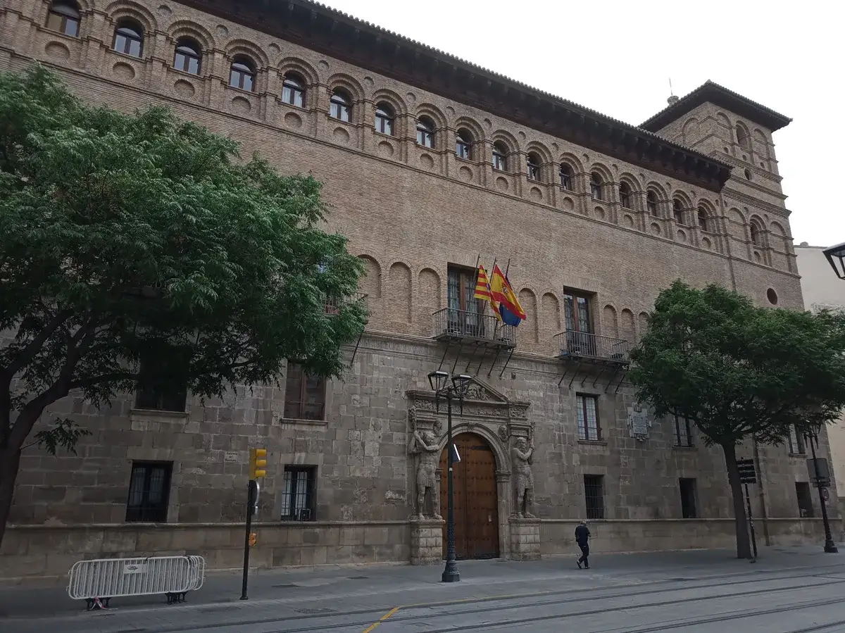 El palacio de los Luna en Zaragoza, con su fachada de ladrillo y piedra, luce imponente. Dos árboles verdes flanquean la entrada principal.