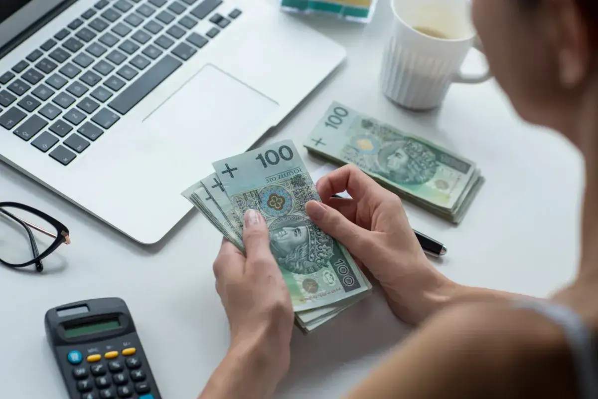 Ręce liczą banknoty 100 zł. Obok laptop, kalkulator i okulary. Zatory płatnicze to problem, który dotyka wielu firm.