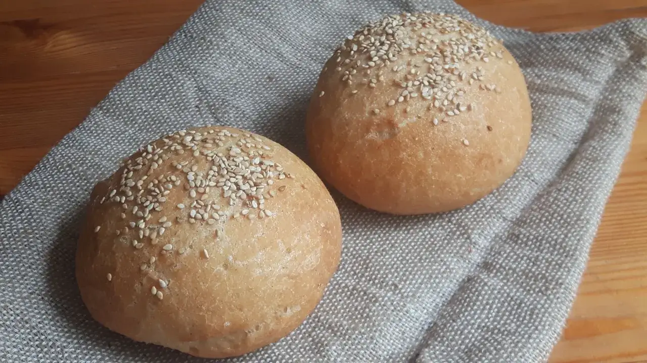 Dos panes sin gluten dorados y esponjosos, cubiertos de semillas de sésamo, descansan sobre una tela rústica.