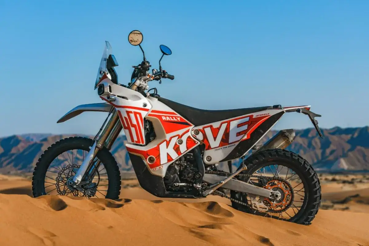 Biało-czerwony motocykl Kove 450 Rally, gotowy na lekkie enduro, stoi na piaszczystej wydmie.