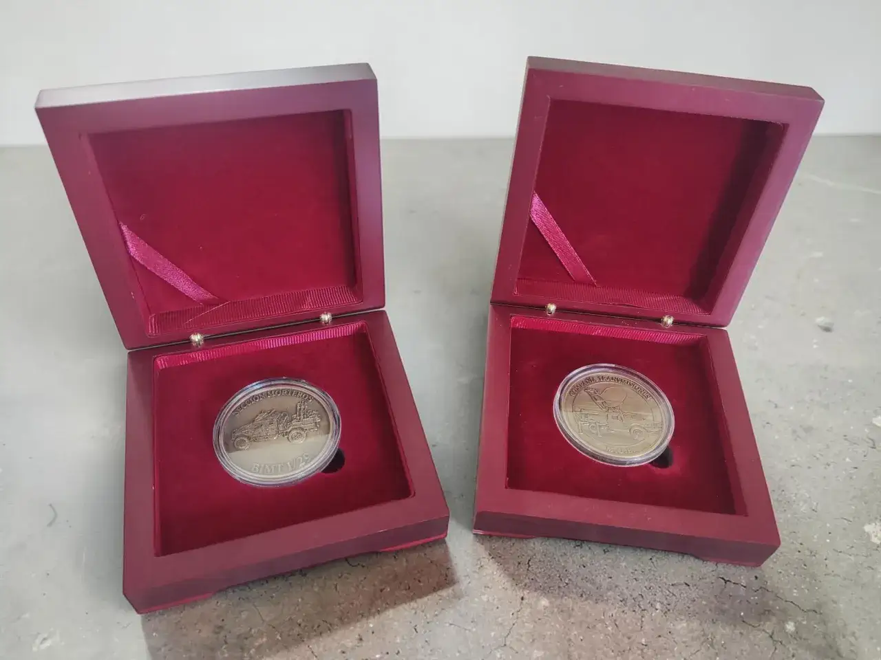 Dos cajas abiertas de terciopelo rojo exhiben dos monedas personalizadas con grabados de vehículos.