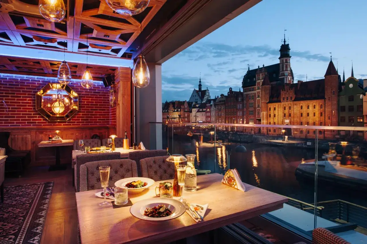 Elegancki wine bar w Gdańsku z widokiem na Motławę. Stoły nakryte, gotowe na wiecz&oacute;r z winem i widokiem na zabytkową zabudowę.
