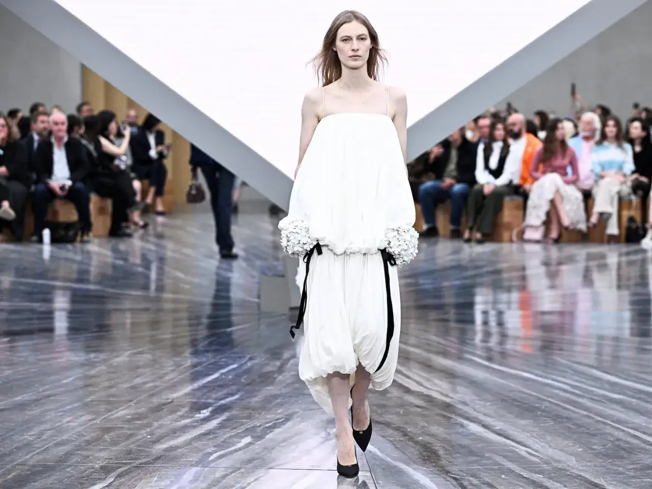 Modelo desfila un vestido blanco voluminoso con detalles florales y cintas negras, una creación de Jonathan Anderson, ante un público atento.