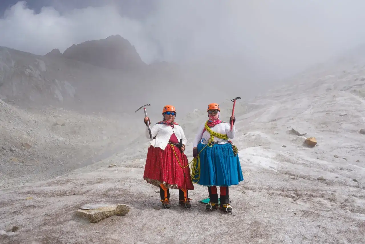 Deux cholitas escaladoras, fières et équipées de piolets, posent sur un glacier brumeux.