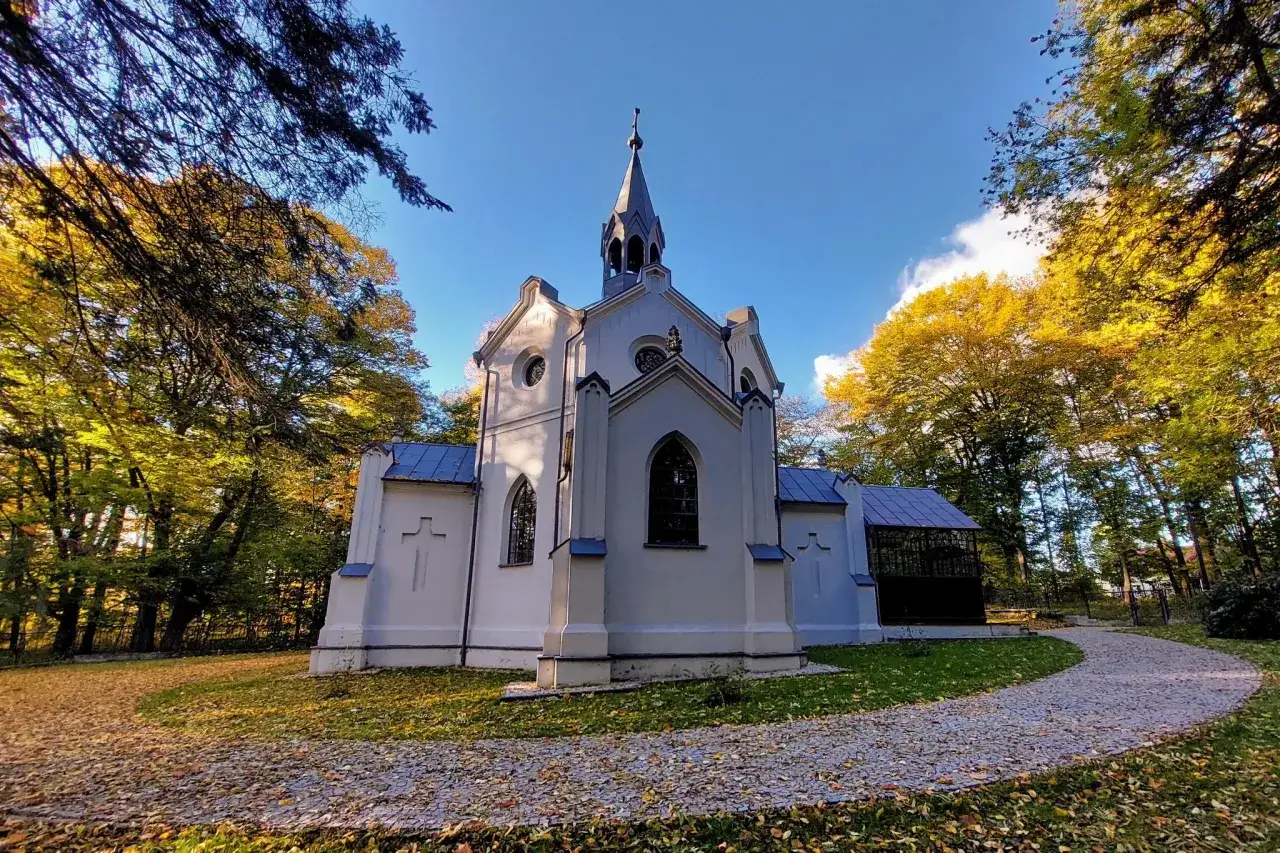 Biała kaplica w otoczeniu jesiennych drzew. Ścieżka wysypana liśćmi prowadzi do niej. W oddali widać konopnicę.
