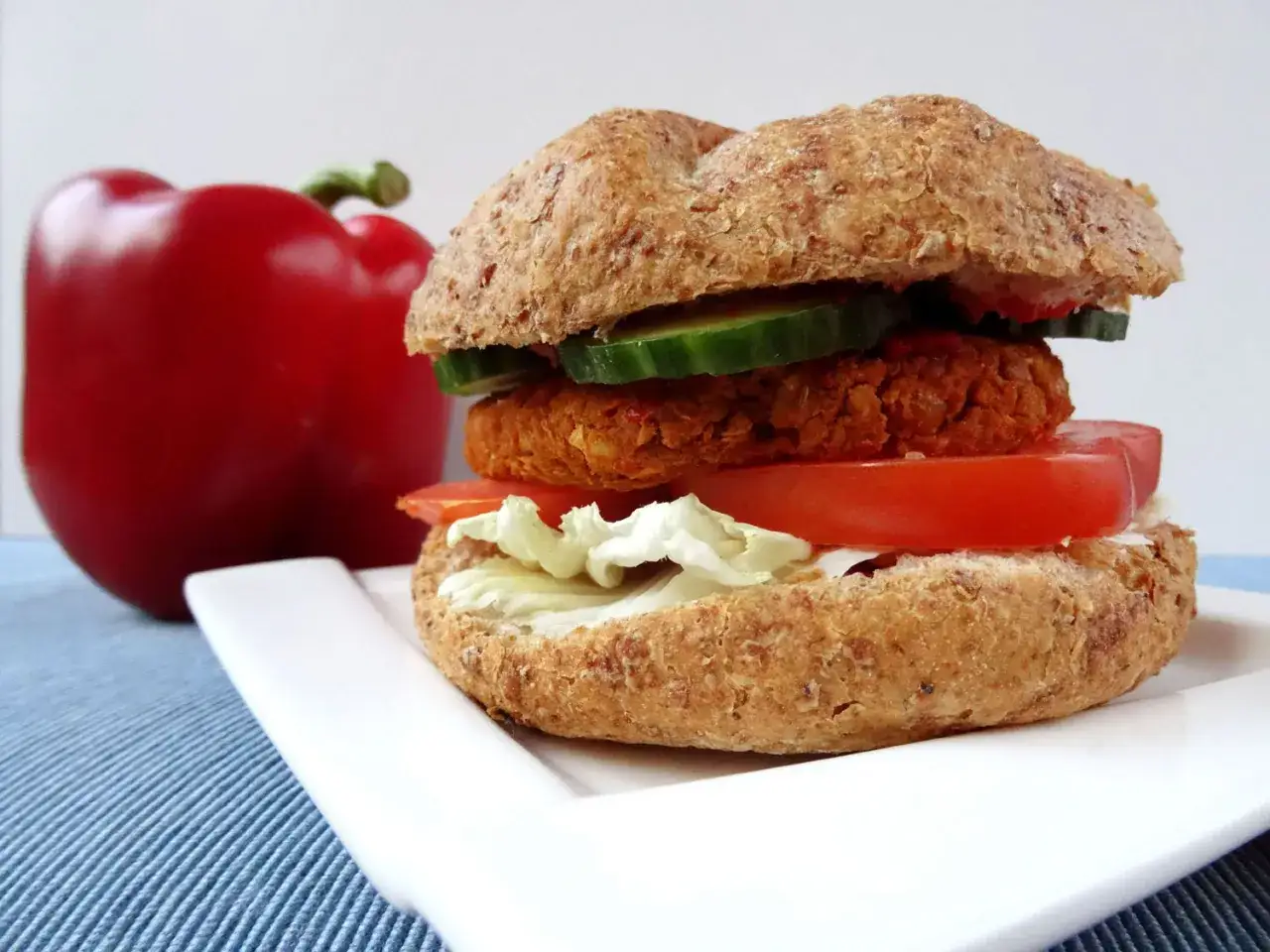 Pyszny burger z ciecierzycy z pomidorem, sałatą i og&oacute;rkiem na białym talerzu, obok czerwona papryka.