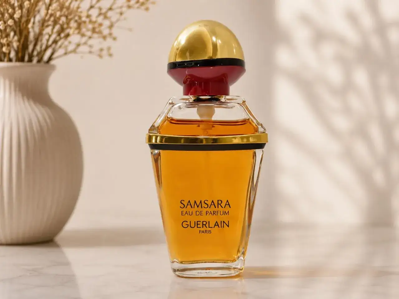 Elegancka butelka Samsara Guerlain z płynem w kolorze bursztynu, obok wazonu z suszonymi kwiatami.
