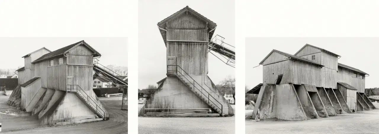 Strutture industriali in legno e cemento, tipiche delle opere di Hilla Becher, con scale esterne e nastri trasportatori.